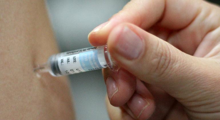 A foto mostra o detalhe de uma mão aplicando uma vacina em um braço. 