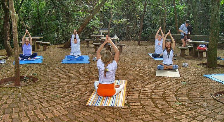 Foto mostra seis pessoas sentadas em tapetes praticando yoga ao ar livre.