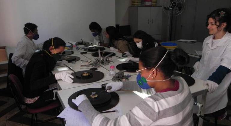 Sete pessoas sentadas em grande mesa com objetos de som e imagem, participando oficina de preservação audiovisual