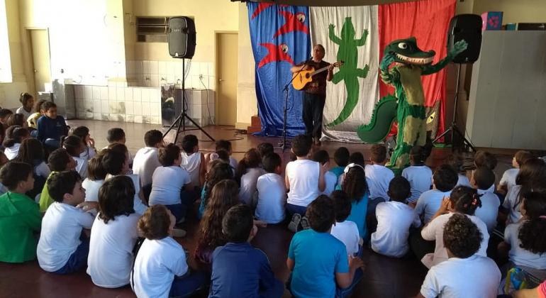 Dezenas de crianças sentadas no chão assistindo apresentação musical/teatral