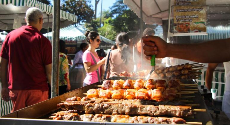 Feirante prepara churrasco na Feira da Afonso Pena. Ao fundo, pessoas e barracas