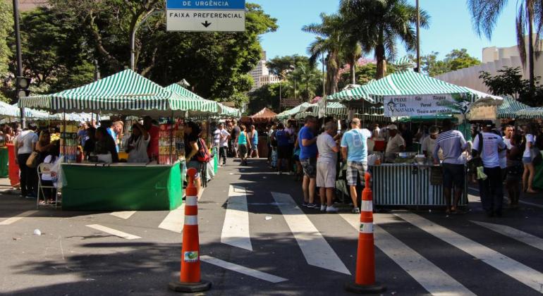 Barracas da Feira montadas na Avenida Afonso Pena. Há uma placa de trânsito escritas "Veículo em Serviço de Urgência - Faixa Preferencial". Ao fundo, à direita e à esquerda, há árvores.