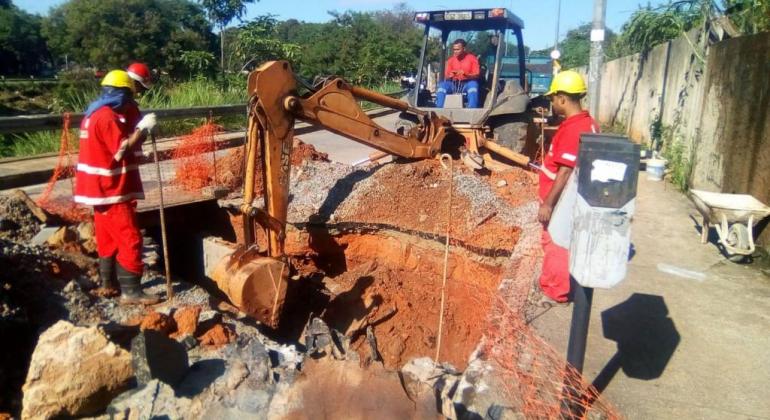 Quatro trabalhadores abrem um buraco no solo com um trator para acessar a rede de drenagem.