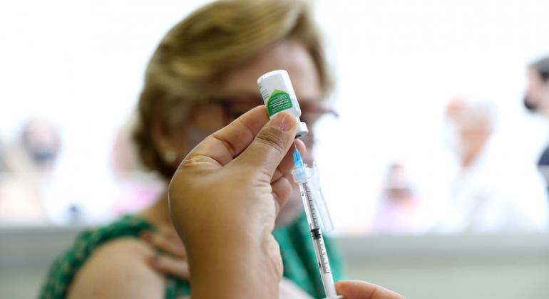 Imagem de seringa e agulha com frasco de vacina contra a gripe. Ao fundo, idosa aguardando a aplicação