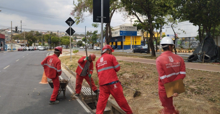 Manutenção no Anel Rodoviário marca gestão da Prefeitura de BH Quatro trabalhadores com uniformes vermelhos e chapéus estão reparando o asfalto em uma rodovia, provavelmente o Novo Anel Rodoviário de Belo Horizonte. Eles usam pás e um balde preto para cobrir buracos e rachaduras no pavimento. Ao fundo, um caminhão basculante amarelo está estacionado, enquanto uma placa de trânsito e uma torre de metal indicam a proximidade de uma via movimentada.