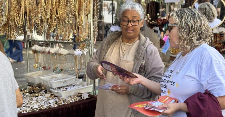 Mulher distribui Leque e panfleto informativo em feira aberta, durante o dia