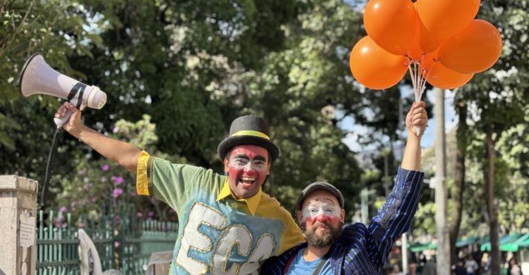 Dois palhaços com pernas de pau, megafone e balões laranja, abraçados, em frente ao Parque Municipal Américo René Giannetti, durante o dia