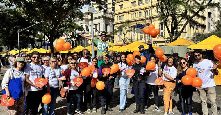 Cenra de 13 pessoas com a camiseta da Campanha Maio Laranja, dois guardas civis municipais e um palhaço com perna de pau, todos segurando balões laranja da campanha, em frente a Feira de Artes e Artesanato da Avenida Afonso Pena, durante o dia