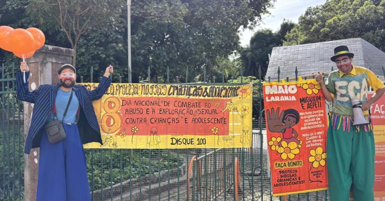 Dois palhaços com pernas de pau com cartazes e balões da Campanha Maio Laranja, em frente ao Parque Municipal Américo René Giannetti, durante o dia