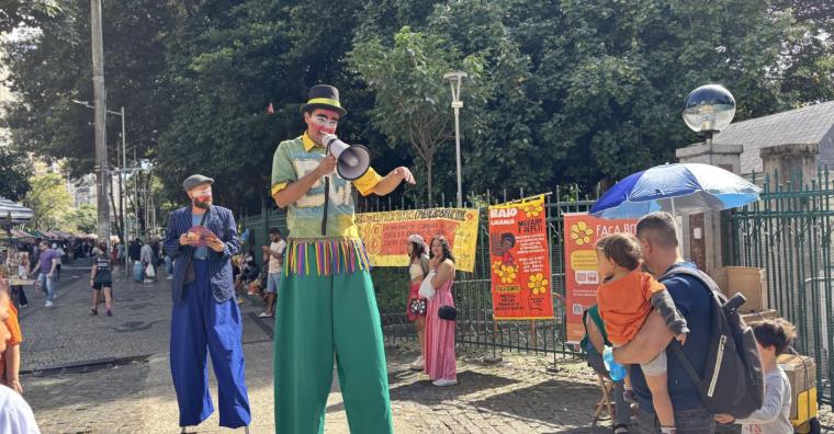 Dois palhaços com pernas de pau com cartazes e balões da Campanha Maio Laranja, abordam os passantes em frente ao Parque Municipal Américo René Giannetti, durante o dia