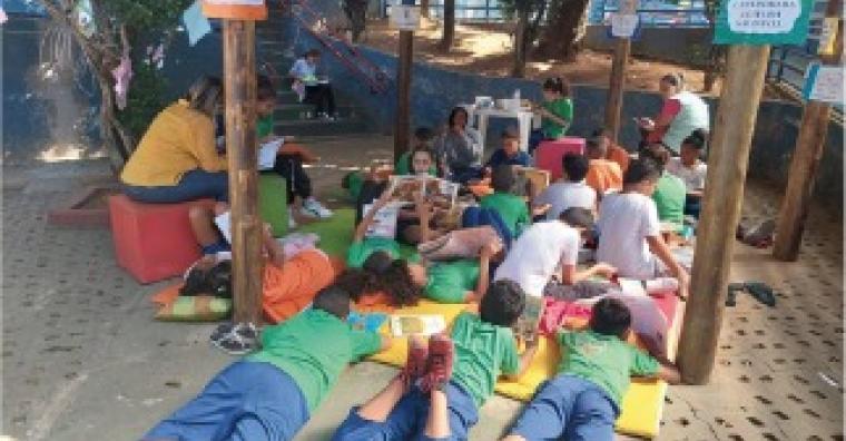 #paratodosverem: Fotografia colorida de estudantes reunidos para ler. Cerca de duas dezenas de crianças estão reunidas em um lugar identificado por uma placa como “Cantinho da Leitura Saudável”, composto por tapetes e bancos coloridos embaixo de uma estrutura de madeira. Algumas estão sentadas e outras deitadas, mas todas possuem algum livro em mãos.