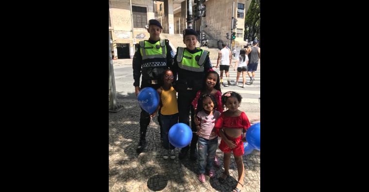 Guardas municipais posam para a foto, abraçados com crianças que seguram balões azuis.