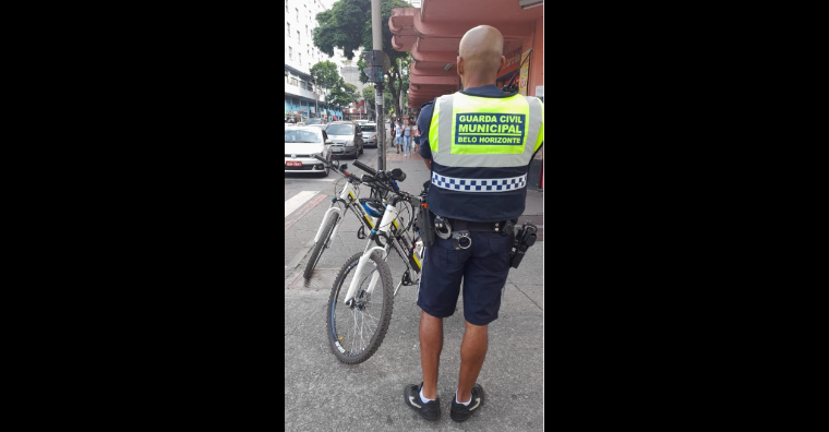 Um homem com uniforme da guarda municipal está de costas, junto a sua bicicleta.