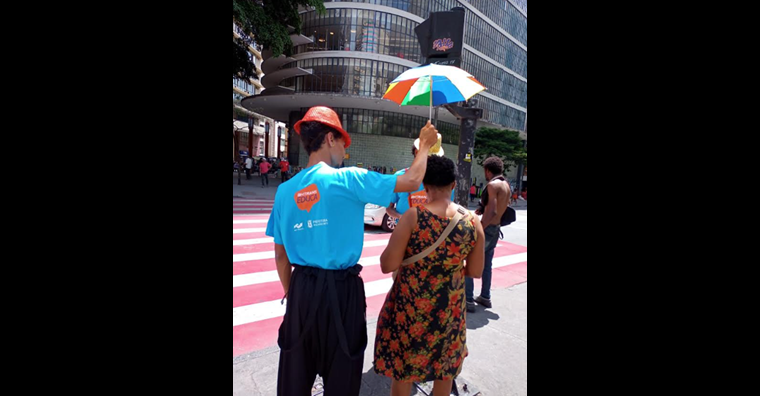 Educador de blusa azul segurando um guarda-sol colorido sob a cabeça de uma senhora que vai atravessar a rua.
