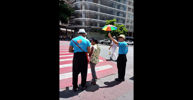 Educador de blusa azul segurando um guarda-sol colorido ao lado de uma senhora que vai atravessar a rua, enquanto outro educador conversa com ela esperando para atravessar a rua.
