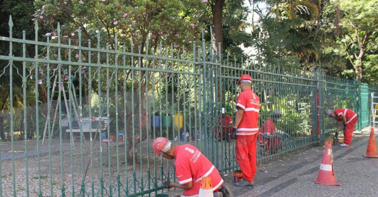 Homens uniformizados com roupa vermelha da PBH trabalham pintando o gradil verde do parque