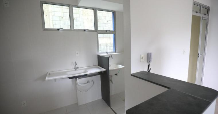 Imagem do interior de uma Unidade Habitacional. O cômodo mostrado é uma cozinha, com paredes brancas, pia branca e uma bancada onde o tampo é preto. Em uma das paredes existe um interfone.