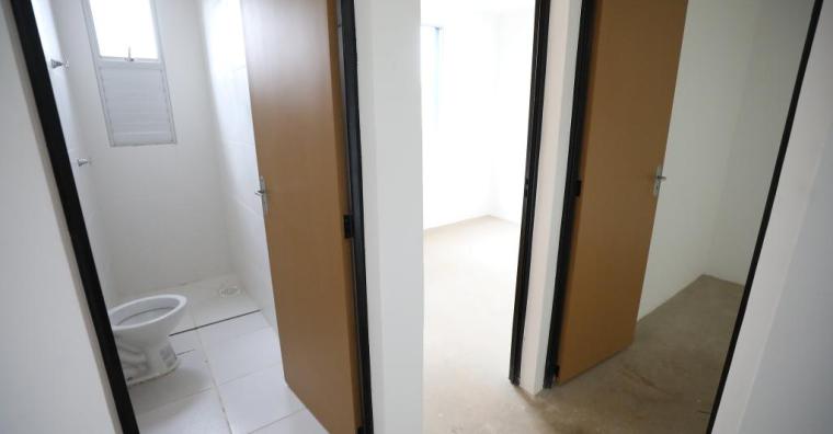Imagem do interior de uma Unidade Habitacional. O cômodo mostrado é um corredor, onde todas as paredes são brancas e as portas de um banheiro e de um quarto estão abertas.