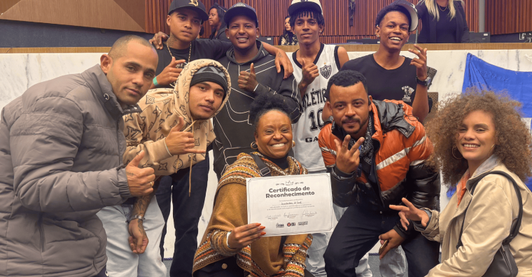 Homenagem na Cãmara Municipal de Belo Horizonte Grupo de jovens e uma mulher segurando um Certificado de Reconhecimento sorriem reunidos em um plenário, fazendo gestos com as mãos.
