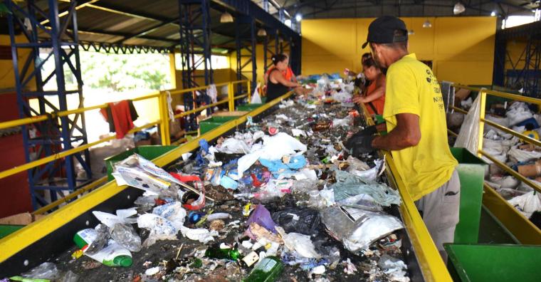 Um catador de materiais recicláveis usando camisa amarela recolhe resíduos que passam por uma esteira automática. Ao fundo, algumas catadoras também realizam a triagem de resíduos recicláveis.