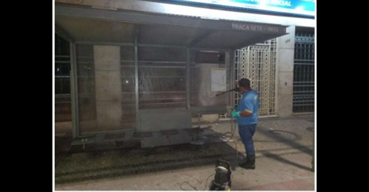 Homem vestindo camisa azul e luvas lavando com máquina o abrigo de parada de ônibus