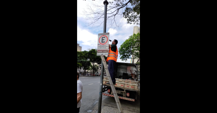 Homem vestindo colete laranja troca sinalização vertical de ônibus durante o dia.