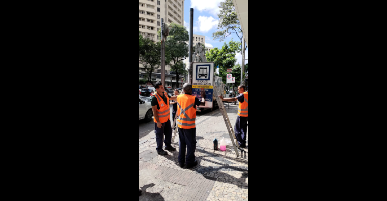 Homens de colete laranja trocam sinalização vertical de ônibus durante o dia.