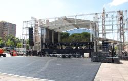 A foto mostra a montagem de um grande palco para um evento ao ar livre, em uma área ampla e pavimentada. A estrutura é composta por treliças metálicas que sustentam a cobertura, os equipamentos de iluminação e os sistemas de som. Nas laterais do palco, há grandes conjuntos de caixas de som, enquanto diversos equipamentos técnicos estão distribuídos na parte frontal e interna da estrutura. No chão, em frente ao palco, há uma extensa área coberta por placas escuras, provavelmente destinada ao público.