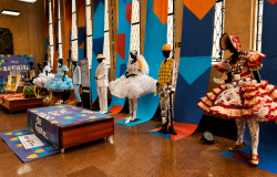 Exposição de figurinos do Arraial de Belô 2026 no hall da Sede da Belotur. Manequins exibem trajes típicos de quadrilhas juninas, com vestidos coloridos, chapéus e roupas ornamentadas. 