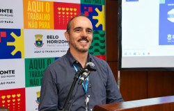 A foto mostra um homem adulto sorrindo, em pé e falando em frente a dois microfones. Ele usa uma camisa azul-marinho, crachá com cordão azul e óculos pendurados na camisa. Ao fundo, há um painel colorido com o brasão e o nome da Prefeitura de Belo Horizonte, além da frase “Aqui o trabalho não para”. À direita, aparece uma tela de apresentação e, na frente, um púlpito de madeira.