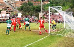 Partida de futebol amador em campo de grama, com jogadores das equipes de uniformes vermelhos e verdes disputando a bola próximo ao gol. Ao fundo, a arquibancada está cheia de torcedores, com casas e vegetação ao redor do campo.