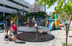 Crianças brincam em uma cama elástica cercada por redes de proteção, enquanto familiares acompanham a diversão. Ao fundo, há brinquedos infláveis coloridos em uma rua arborizada, com prédios ao redor, durante as atividades do programa A Rua é Nossa, da PBH.