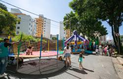 Em uma rua fechada ao trânsito, crianças e famílias participam de atividades recreativas ao ar livre. À esquerda, duas crianças brincam em uma cama elástica cercada por uma rede colorida, acompanhadas por um monitor. Ao centro e ao fundo, há grandes brinquedos infláveis em formatos de polvo, dinossauro e melancia, com várias pessoas reunidas ao redor. Uma mulher caminha pela rua de mãos dadas com uma criança. A cena acontece em um dia ensolarado, cercada por árvores e prédios residenciais.