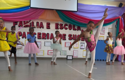 #paratodosverem: crianças se apresentam em um palco escolar decorado com tecido colorido nas cores amarelo, azul, rosa e verde. No fundo, está a frase “Família e Escola, Parceria de Sucesso” e um homem em pé, à esquerda. As crianças usam roupas como vestidos, collants, saias de balé e bermudas. Elas realizam movimentos de dança e ginástica. À direita, pessoas adultas assistem sentadas e aplaudem. 