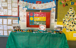 #paratodosverem: a foto mostra murais coloridos sobre alimentação saudável e sustentabilidade com trabalhos das crianças. Ao centro, há um painel com sinalização de cores – vermelho (Evite!), amarelo (Modere!) e verde (Prefira!) – e figuras de alimentos coladas em cada faixa. Em frente, uma mesa coberta com toalha verde exibe pratos com alimentos de brinquedo.