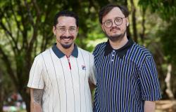 Dois homens brancos posam lado a lado em uma área arborizada, sorrindo para a foto. Gu da Cei veste camisa pólo branca com listras finas e gola azul-marinho; Benedito Ferreira está de camisa azul-marinho com listras verticais brancas e azuis. Ambos usam óculos.