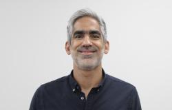 Homem de meia-idade aparece em um retrato de frente, do peito para cima. Ele tem cabelo curto grisalho, sobrancelhas grossas e barba curta também grisalha. Está sorrindo levemente para a câmera. Veste uma camisa preta de botões. O fundo da imagem é liso e claro, sem outros elementos.