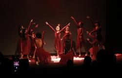 Grupo de dançarinas no palco, vestindo vestidos vermelhos, em uma apresentação. Uma bailarina central está com os braços erguidos em forma de V, rodeada por outras que espelham sua pose. A iluminação é dramática, com foco nas bailarinas e o restante do palco escuro. 