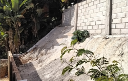 Estrutura de cimento cinza claro feita para contenção de encosta com risco de desabamento, com plantas de médio e grande porte ao fundo. 