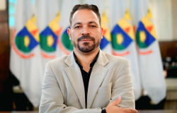 Homem branco de braços cruzados, usando camisa preta e blazer bege