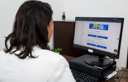 Mulher com uma blusa branca sentada em frente ao computador com o portal de serviços da PBH na tela. 