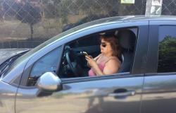 Motorista feminina, com carro estacionado e janela aberta, confere aplicativo no celular, durante o dia.