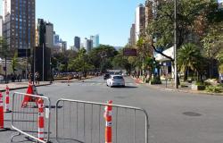 Avenida com cones e grades de interdição e carros passsando ao fundo, durante o dia. 