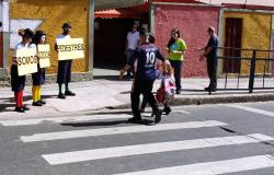 Três pessoas, vestidas de palhaço, na calçada, em frente a um local de travessia de pedestres, com três placas que formam o dizer: "somos todos pedestres". Na travessia, um adulto atravessa com uma criança. 