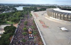 Bloco de rua com mais de quinhentas pessoas em avenida, ao lado do Mineirão, tendo ao fundo o Mineirinho e a Lagoa da Pampulha, durante o dia. 