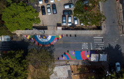 Imagem aérea de uma rua com pinturas e um estacionamento de carros ao lado, durante o dia. 