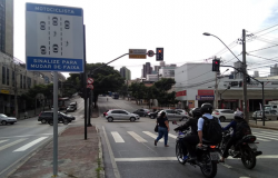 Motociclistas em avenida, ao lado de placa, onde está escrito: "Motociclista: sinalize para mudar de faixa". 