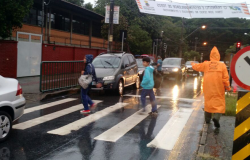 Agente de trânsito com capa de chuva acompanha alunos que atravessam a faixa de pedestre em direção à escola, durante o dia. 