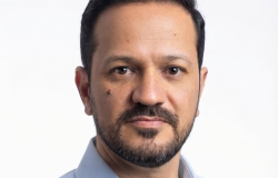 Homem adulto, de cabelos curtos escuros e barba aparada, usando camisa social azul-clara. Está de frente para a câmera, com expressão séria e fundo branco, em um retrato com aparência profissional.