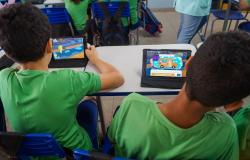 Dois estudantes de uniforme verde e azul, sentados de costas para a câmera, usam tablets em uma sala de aula de uma escola no Brasil. O tablet do aluno da esquerda exibe o jogo de aventura de pular em poças 'Pega-poça', e o do aluno da direita mostra o jogo da escola. Os alunos estão cercados por outros estudantes e cadeiras e mesas brancas e azuis.